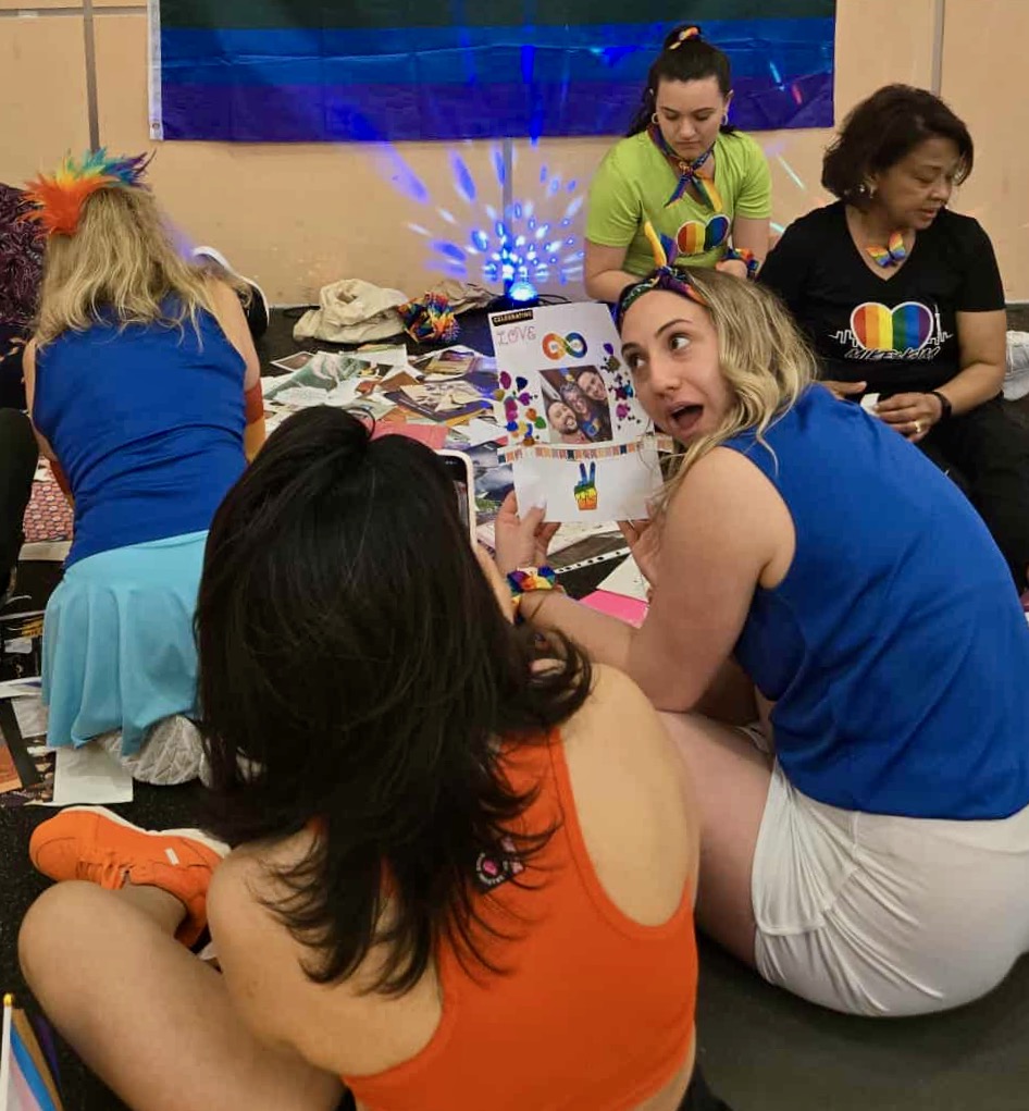 Five women are sitting on a dance stage with collage materials in the middle. In the lower right of the photo, a young woman with a expression happy shows off her collage a friend on her left, who is taking a photo of her.